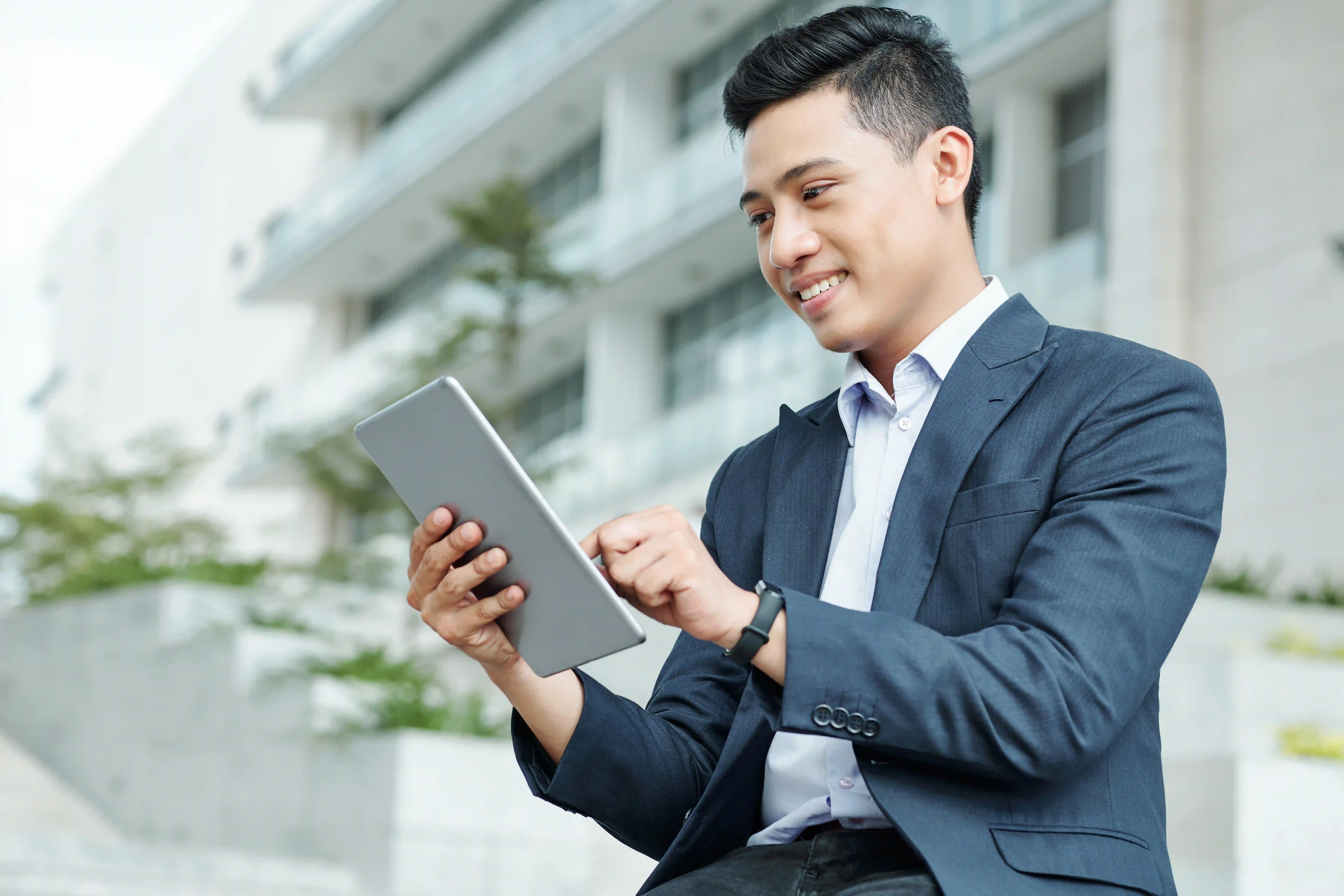Young man in suit uses tablet outside a modern building, smiling.