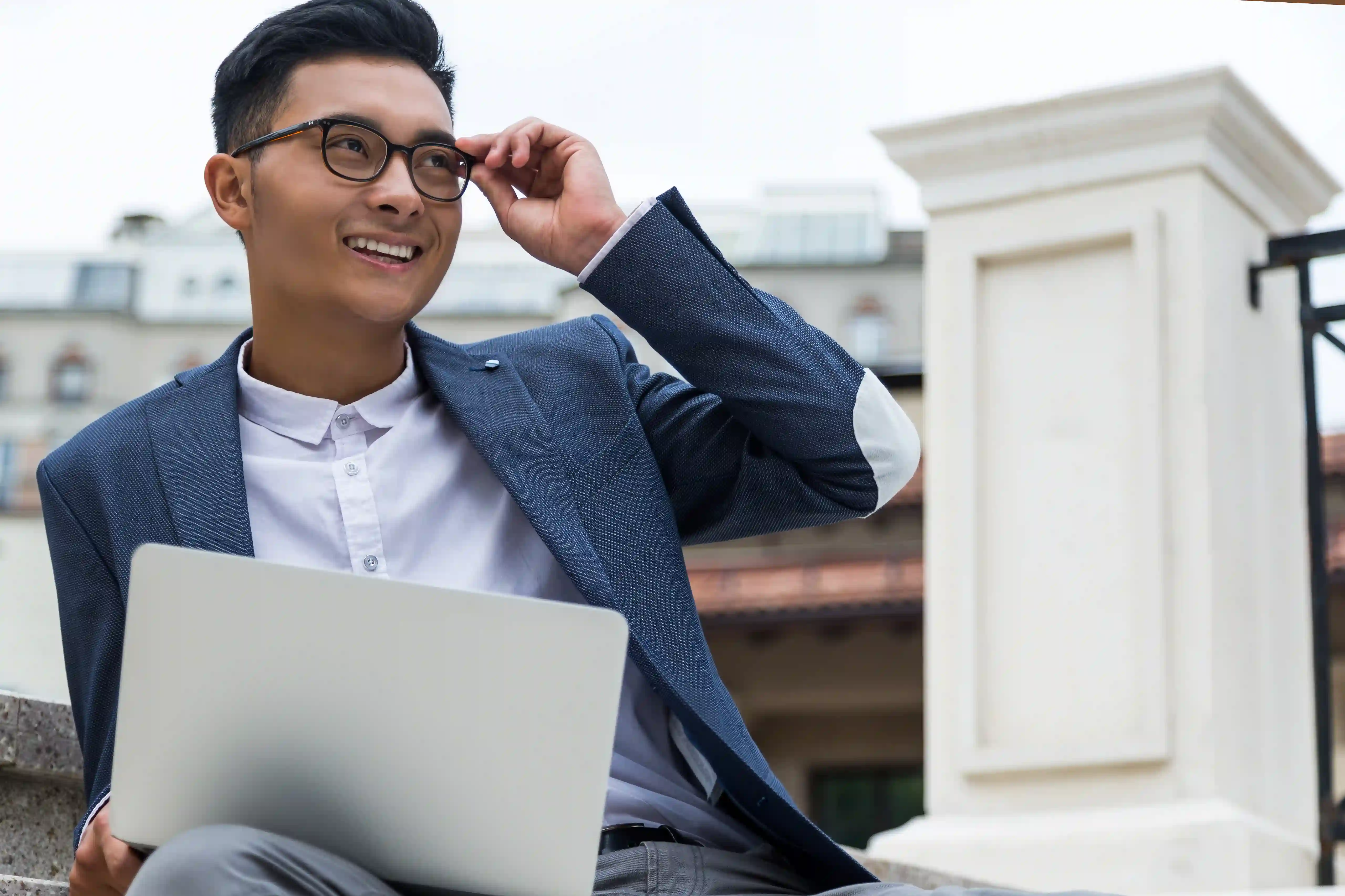 Smiling person with laptop sits outdoors in a bright urban setting.