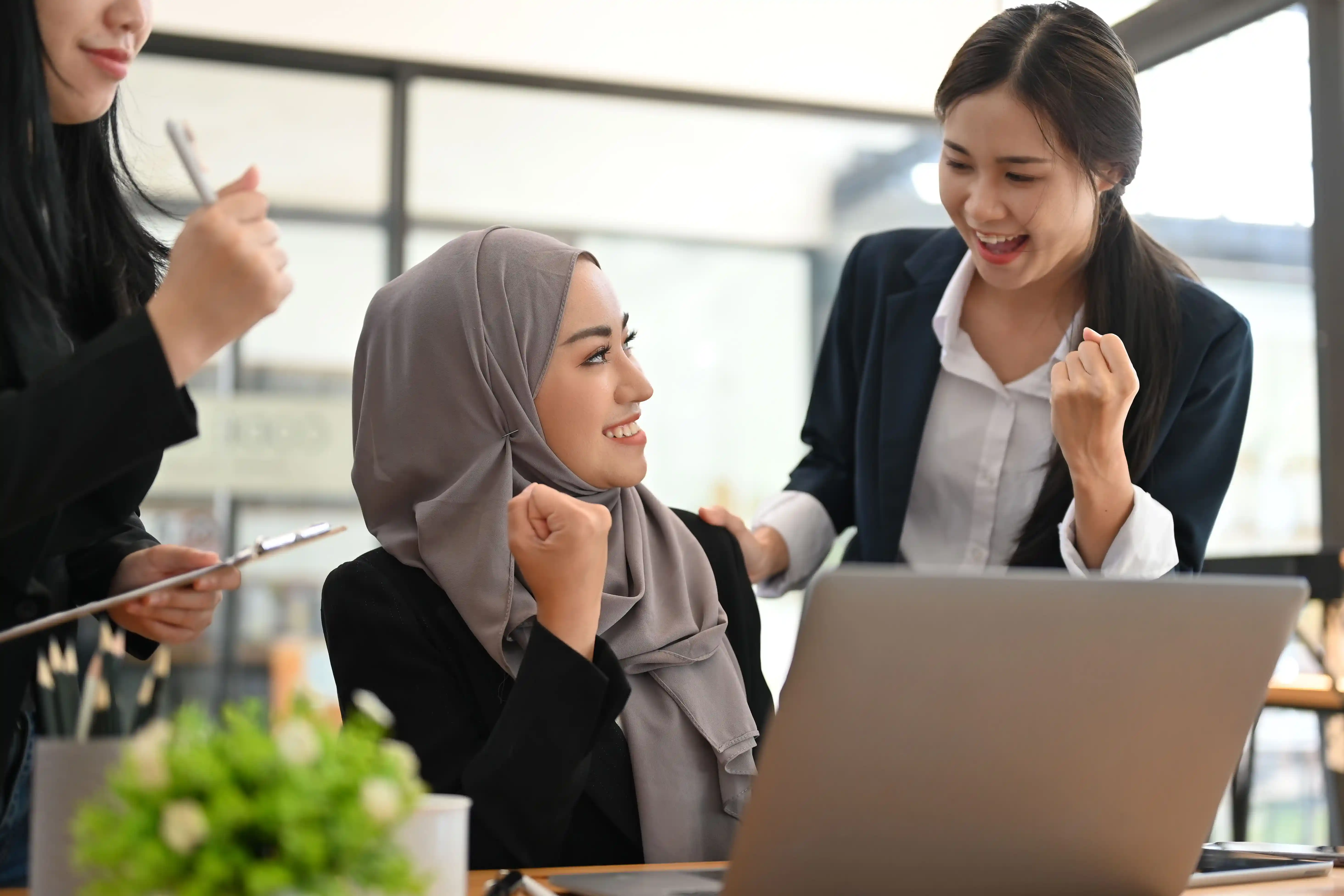 Tiga wanita meraikan kejayaan di pejabat bersama komputer riba dan pokok.