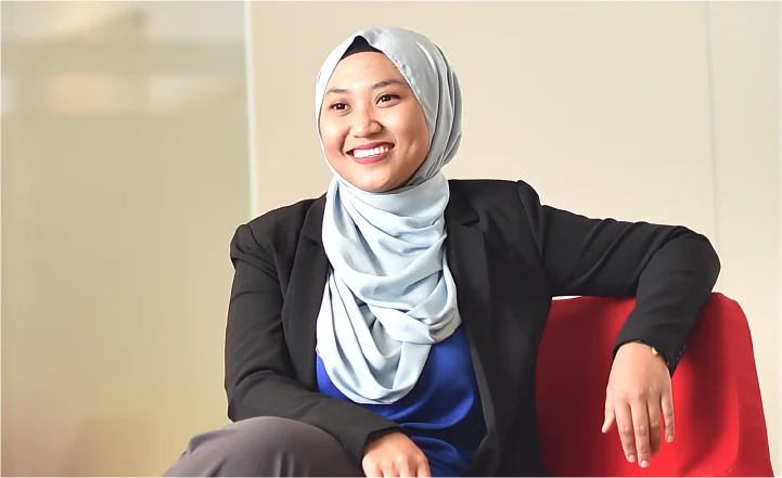 A smiling professional woman in hijab and blazer seated confidently in an office