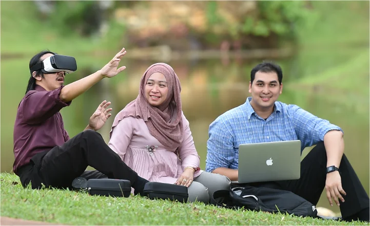 Three colleagues relaxing on grass outdoors, using VR headset and laptop in a park