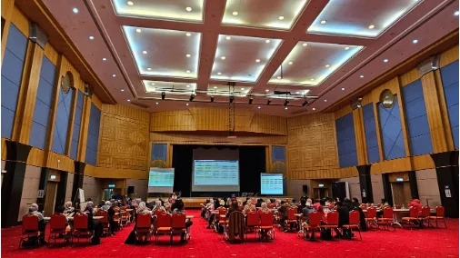 Large event hall with red carpet, round tables and a lit-up stage with dual screens