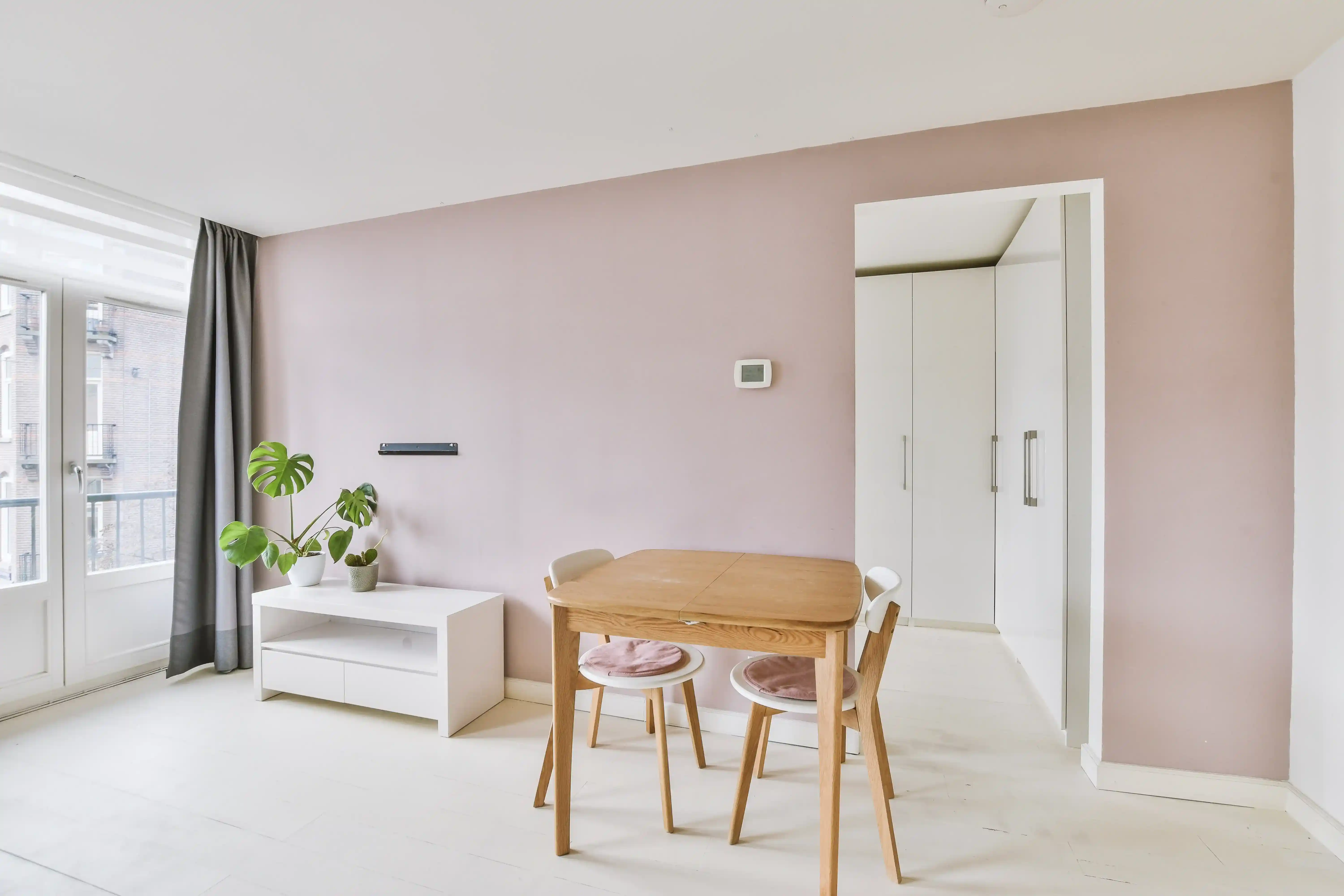 Minimalist dining area with wooden table, chairs, plant, and pastel pink wall.