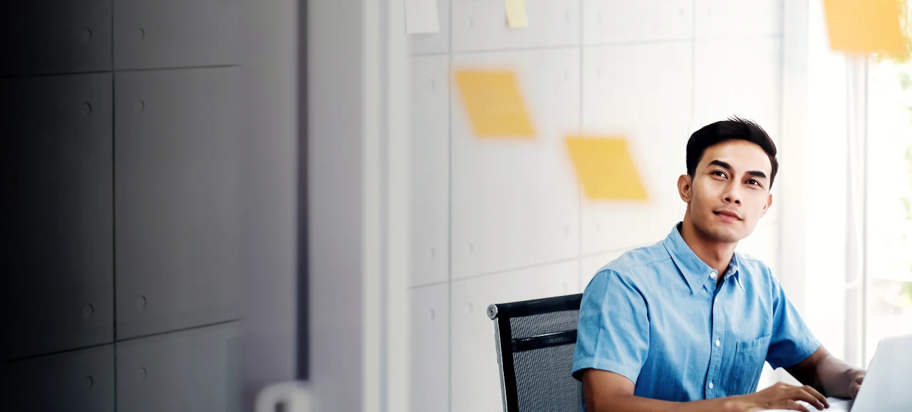 Man in blue shirt works on laptop at desk with sticky notes behind.