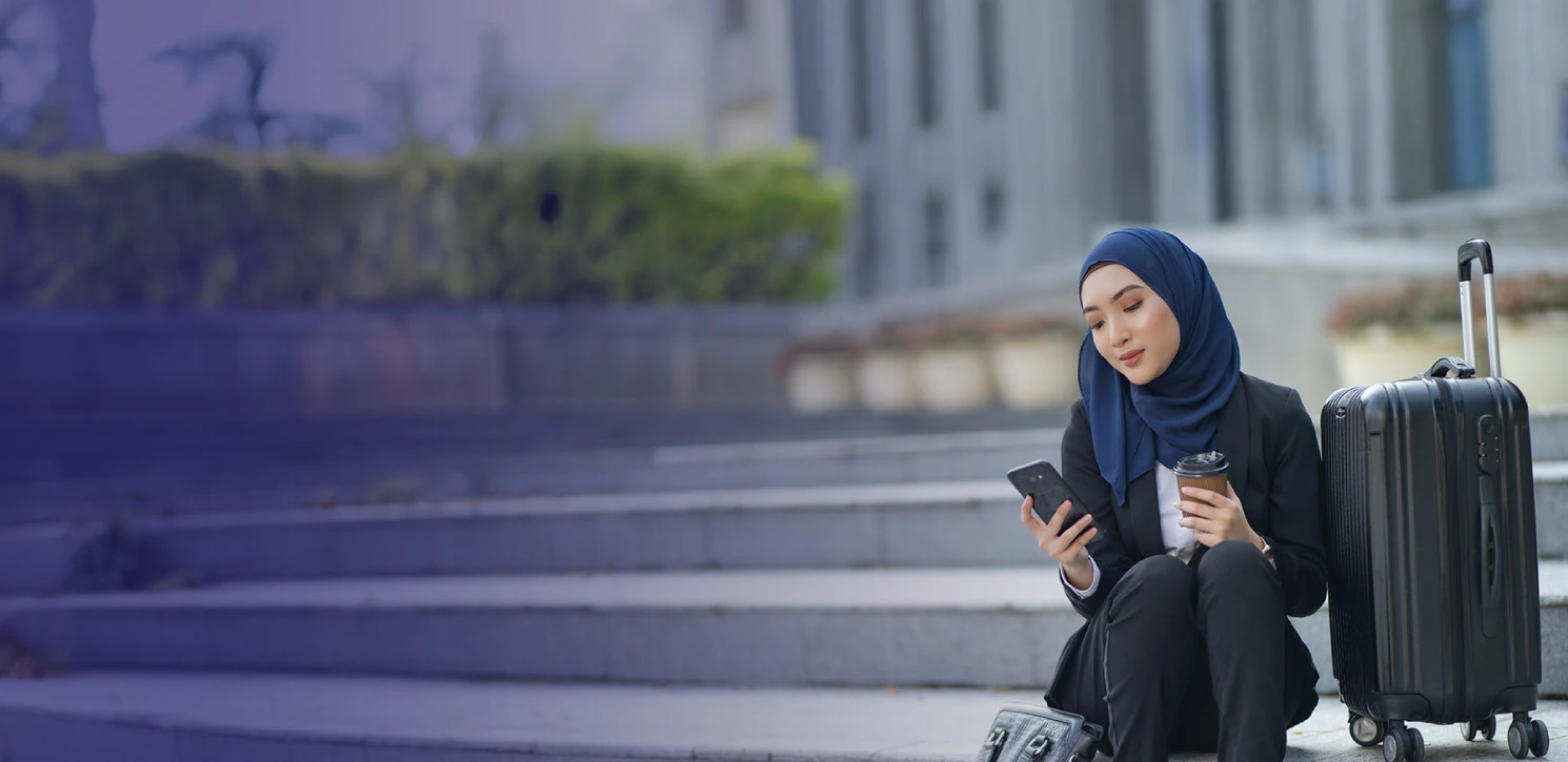 Wanita berhijab biru duduk bersama beg pakaian sambil memegang telefon dan kopi.