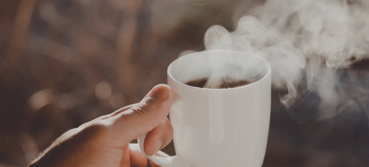 Hand holds steaming coffee mug in a warm, cozy blurred background.